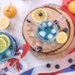 A pitcher and glass of blue lemonade with ice, lemon slices, and blueberries, surrounded by festive decor—this refreshing Fourth of July Drink is the perfect patriotic mocktail for your celebration.
