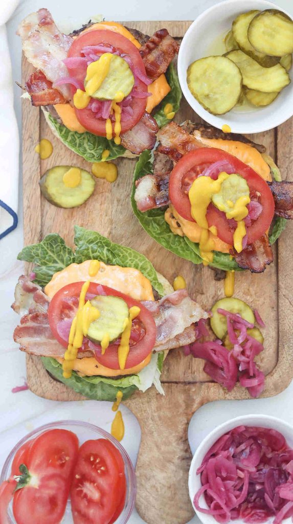 Three lettuce-wrapped Bacon Cheeseburgers topped with bacon, tomato slices, pickles, onions, cheese, and mustard sit on a wooden board. Bowls of pickles, sliced tomatoes, and pickled onions are nearby—a true Best Burger experience.