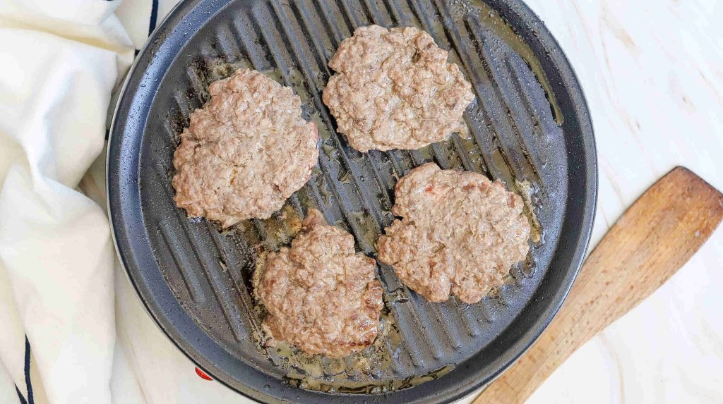 Four irregularly shaped hamburger patties sizzle on a ridged grill pan, perfect for your next American burger recipe, with a wooden spatula and a white cloth nearby.