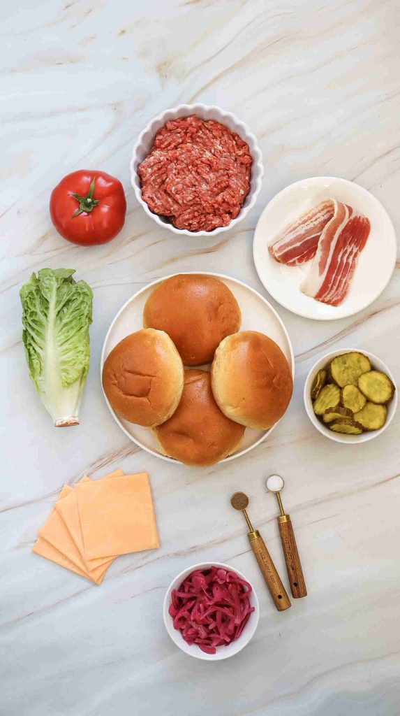 Burger ingredients arranged on a light surface: ground beef, bacon strips, a tomato, romaine lettuce, sandwich buns, sliced pickles, cheddar cheese slices, pickled onions, and small containers of salt and pepper—perfect for the Best Burger or classic Bacon Cheeseburger.