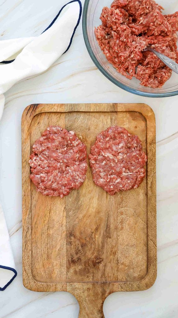 Two raw ground beef burger patties on a wooden cutting board, with a bowl of uncooked ground beef mixture and a white cloth nearby—perfect for crafting your favorite American Burger or trying a new burger recipe.