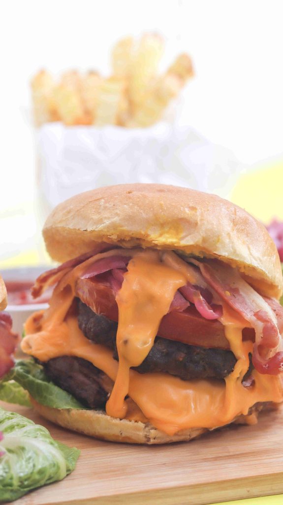 A close-up of a classic American burger piled high with melted cheese, crispy bacon, lettuce, tomato, and a juicy beef patty on a bun, with a side of fries blurred in the background.