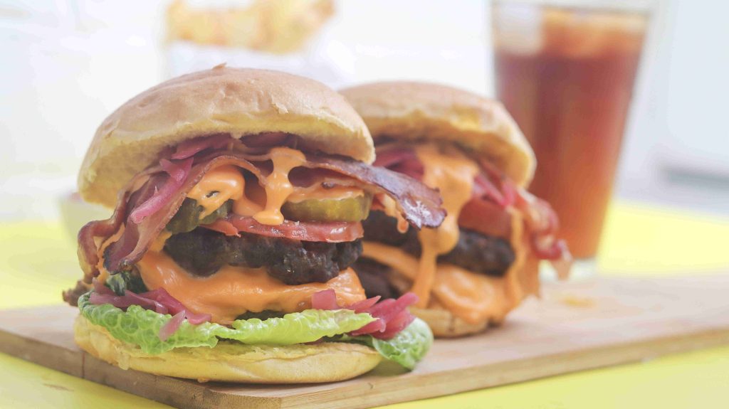 Two loaded bacon cheeseburgers with lettuce, pickles, tomato, melted cheese, and sauce sit on a wooden board—an American burger recipe served with fries and iced tea blurred in the background.