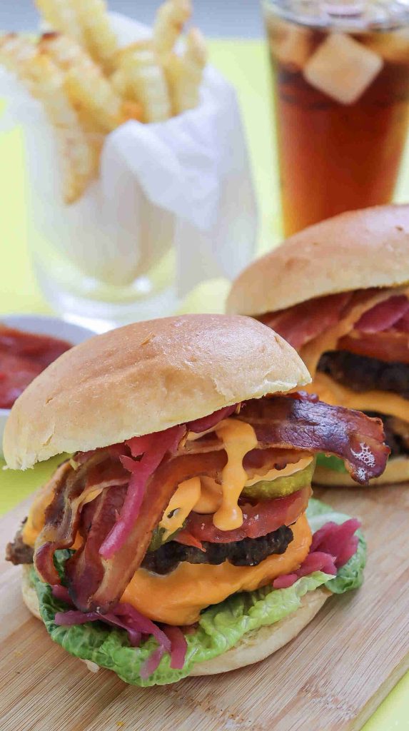 A close-up of two classic American Burgers with lettuce, pickles, tomatoes, onions, and sauce on buns, served on a wooden board. In the background are crinkle-cut fries in a cup and a glass of iced drink.