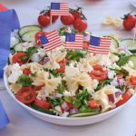 A festive 4th of July pasta salad with tomatoes, cucumbers, onions, herbs, and feta cheese, topped with small American flags. Cherry tomatoes and a wooden spoon with feta sit nearby alongside blue and red streamers for a perfect red white blue side dish.