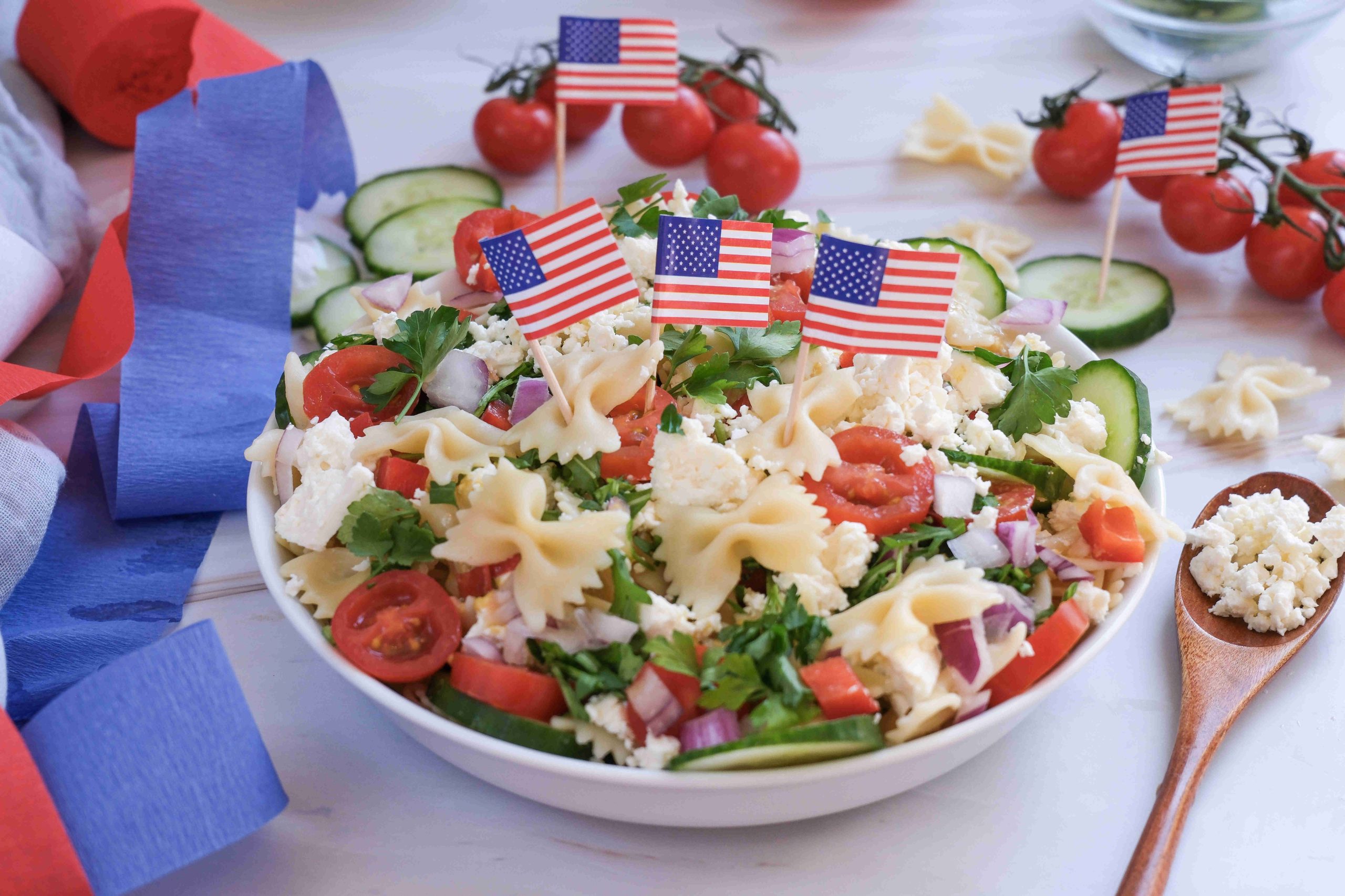 A festive 4th of July pasta salad with tomatoes, cucumbers, onions, herbs, and feta cheese, topped with small American flags. Cherry tomatoes and a wooden spoon with feta sit nearby alongside blue and red streamers for a perfect red white blue side dish.
