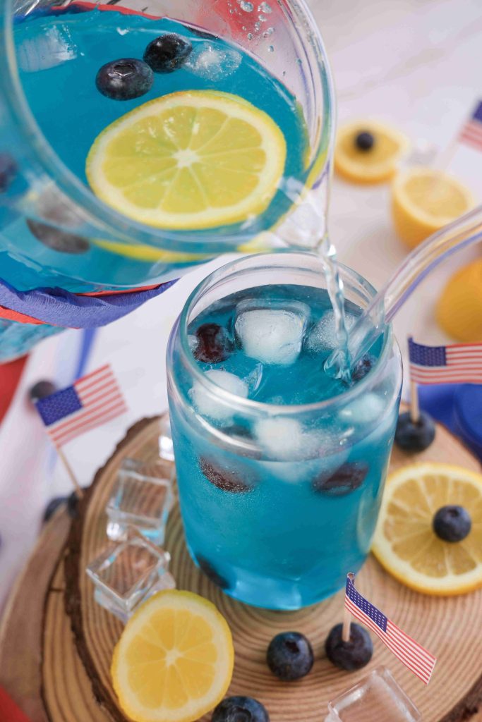 A glass of Sparkling Blue Lemonade with ice, blueberries, and lemon slices is being poured from a pitcher. The drink sits on a wooden surface with lemon slices, blueberries, mini American flags, and clear ice cubes around it—perfect for a Fourth of July Drink.