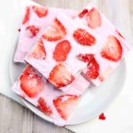 A plate stacked with pink strawberry yogurt bark bars topped with fresh strawberry slices sits on a white cloth decorated with small red hearts. The bars have a creamy texture and a fruity appearance.