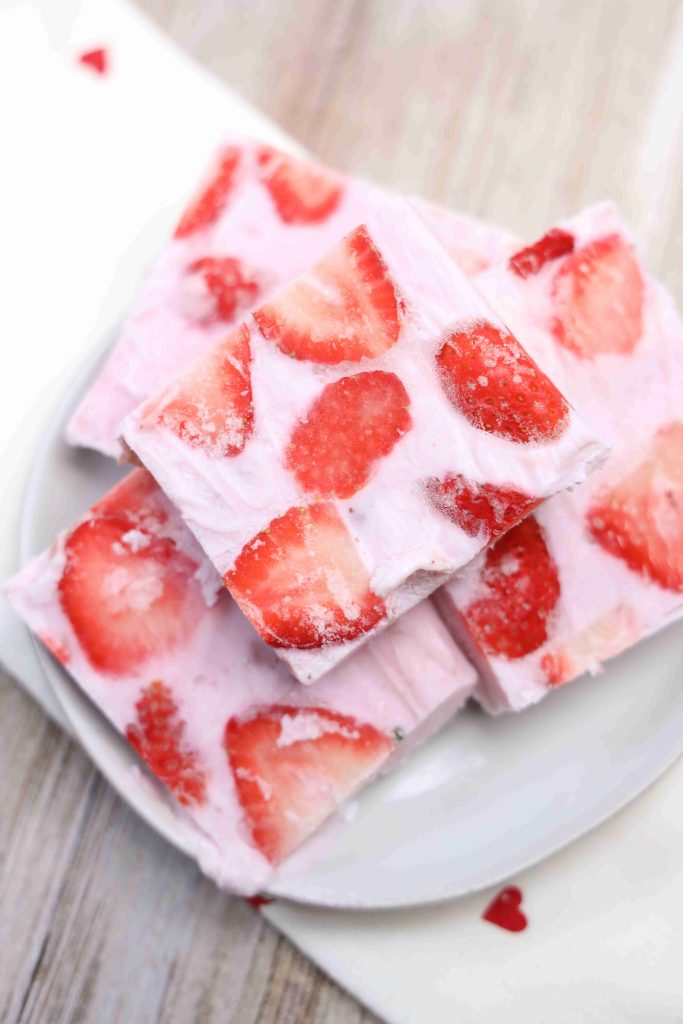 Three pieces of frozen yogurt bark with sliced strawberries are stacked on a white plate, set on a light wooden surface. The bars have a creamy pink color and visible strawberry pieces throughout.
