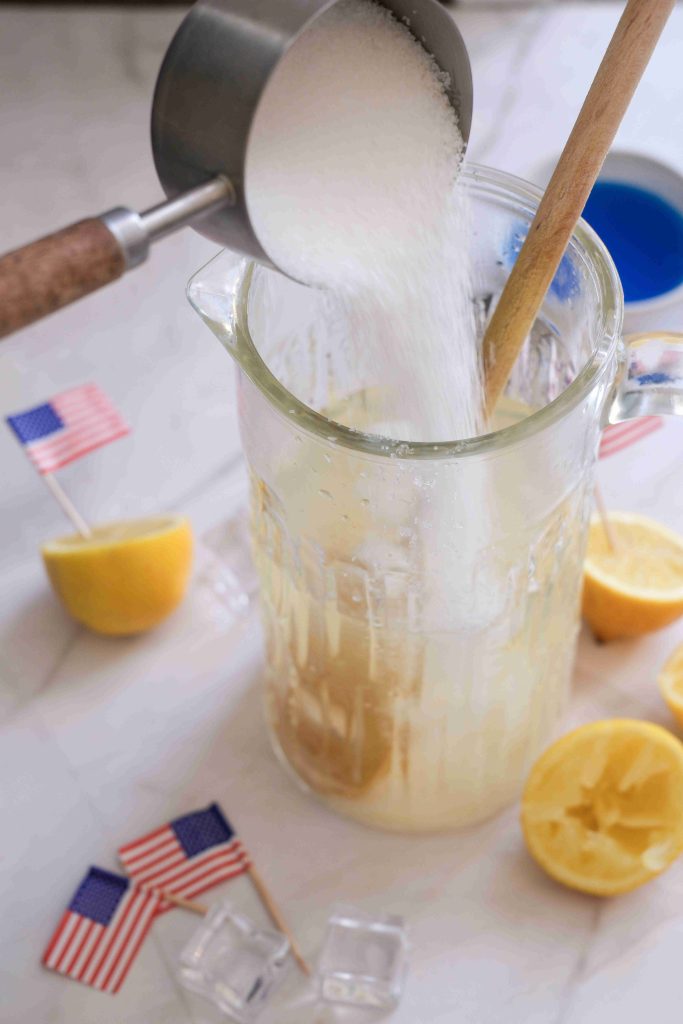 A metal cup pours sugar into a glass pitcher of blue lemonade with a wooden spoon. Squeezed lemons, lemon halves, ice cubes, and small American flags are nearby—perfect for a festive Fourth of July drink or summer mocktail.