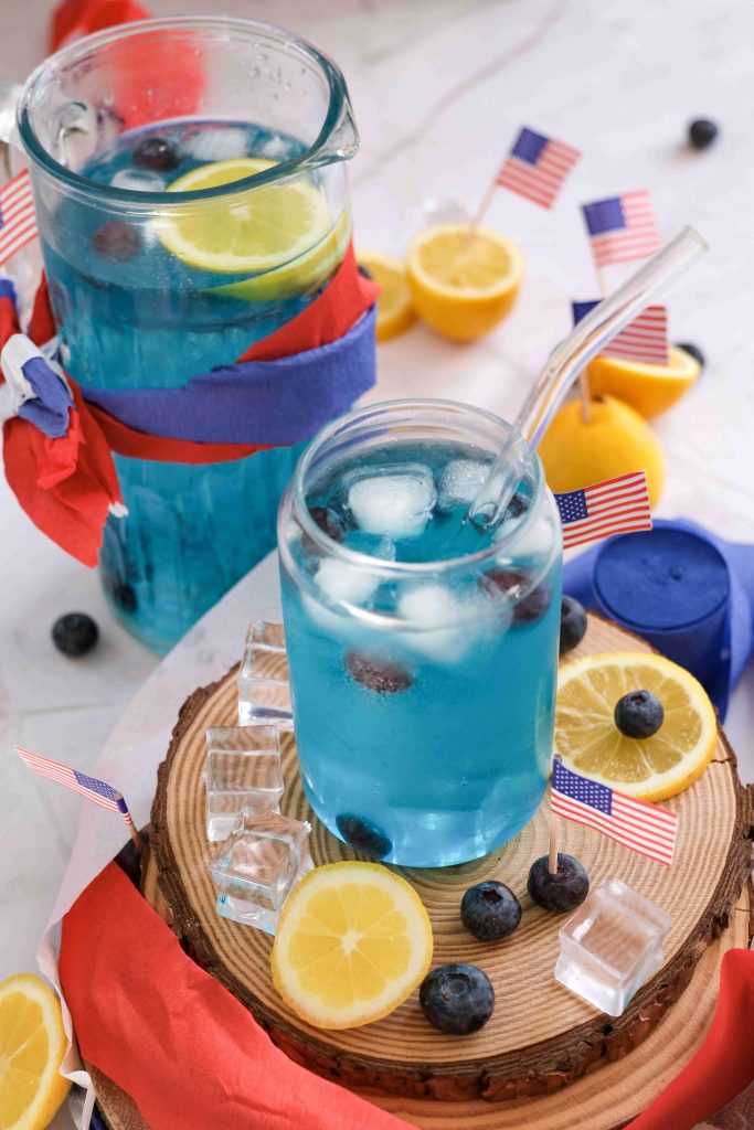 A glass and a pitcher of blue lemonade mocktail with ice, blueberries, lemon slices, and small American flags, surrounded by lemons, blueberries, ice cubes, and red, white, and blue decorations—a perfect Fourth of July drink.