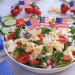 A bowl of 4th of July pasta salad with bow-tie pasta, cherry tomatoes, cucumbers, red onion, cheese, and parsley is topped with small American flags—a festive red white blue side dish perfect for your patriotic celebration.