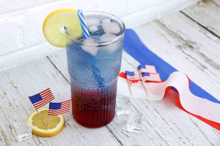 A layered red white and blue drink with ice and a lemon slice sits on a rustic white table, surrounded by mini American flags, lemon slices, ice cubes, and a red, white, and blue ribbon—perfect for patriotic punch at summer BBQs.