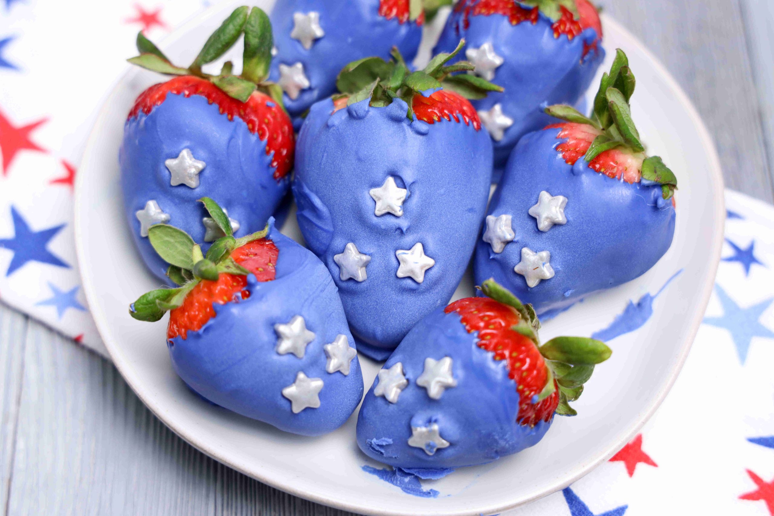 A plate of 4th of July strawberries dipped in blue chocolate and topped with white star-shaped candies. Some red tops peek out, and the plate rests on a star-patterned cloth—making this a festive, foolproof recipe for any celebration.