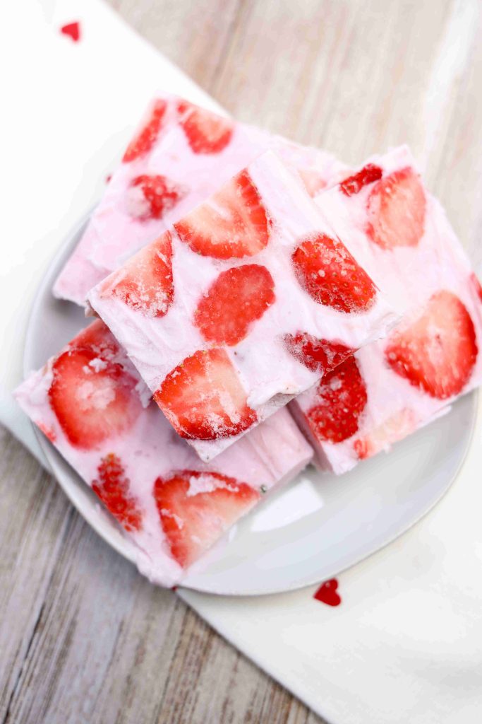 A plate with several squares of yogurt bark topped with sliced strawberries, placed on a white napkin with small red hearts, on a wooden surface.