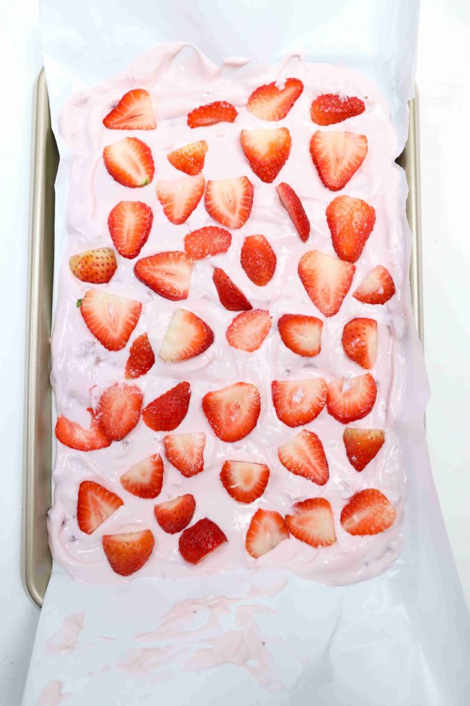 A baking tray lined with parchment paper holds a pink creamy yogurt bark mixture, topped with evenly spaced, sliced fresh strawberries.