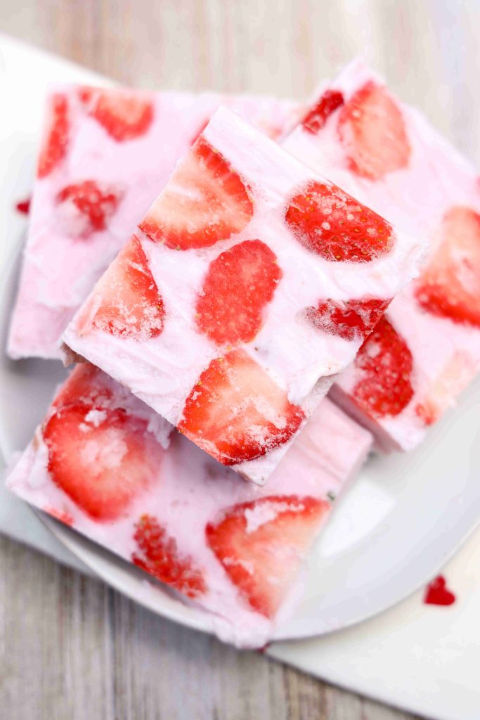 Three squares of yogurt bark with visible strawberry slices are stacked on a white plate. The light pink yogurt and bright red strawberries create a fresh and fruity appearance, making this frozen treat as appealing as it is delicious.