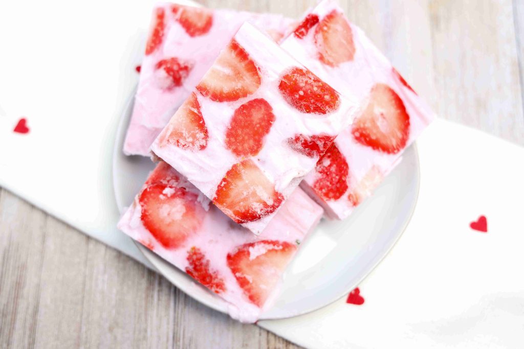 Four squares of pink strawberry yogurt bark with visible strawberry slices, stacked on a white plate. The creamy yogurt bark sits on a white napkin with small red hearts, atop a light wooden surface.