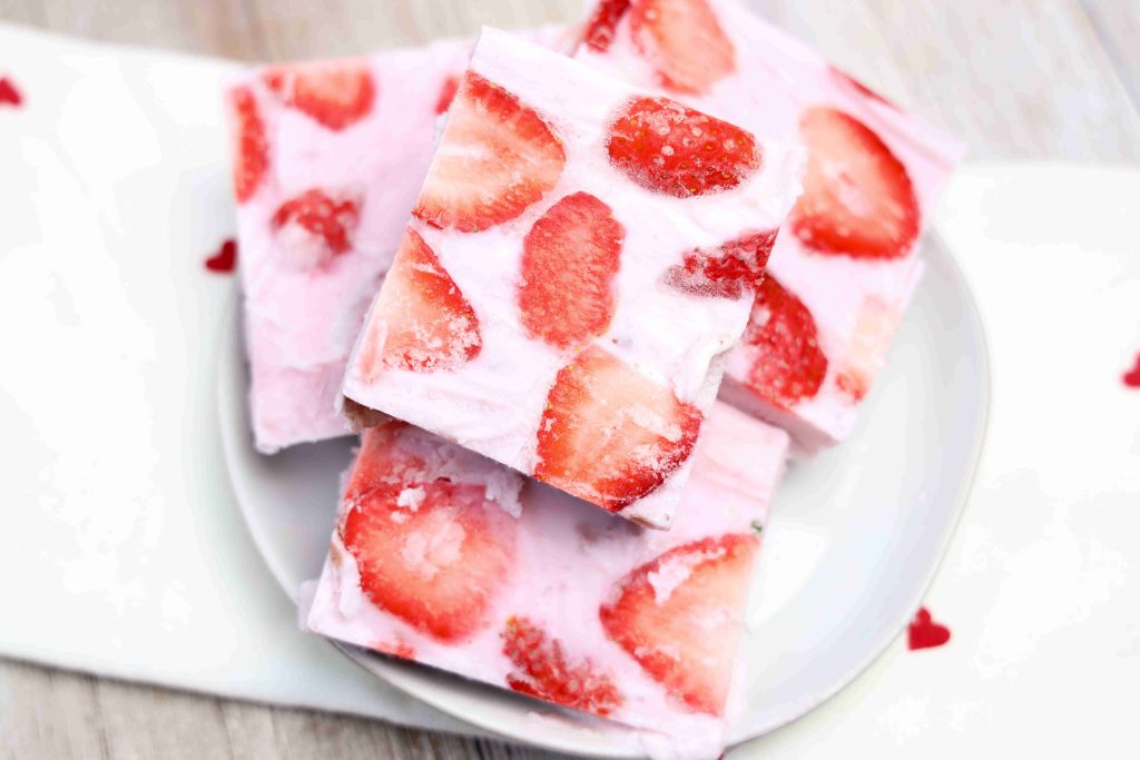 Three square pieces of yogurt bark with visible strawberry slices are stacked on a white plate, placed on a white surface with small red heart decorations.