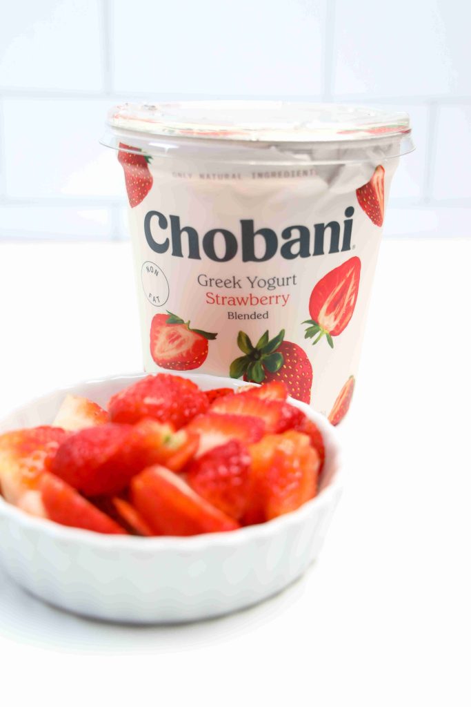 A container of Chobani Greek Yogurt Strawberry flavor sits behind a small white bowl filled with sliced fresh strawberries, perfect ingredients for making refreshing yogurt bark, on a white surface.