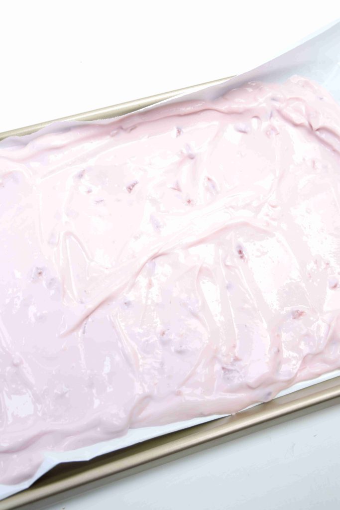 A baking sheet lined with parchment paper is covered in a thick, pale pink yogurt bark mixture with small chunks, likely a dessert base, spread evenly across the surface.