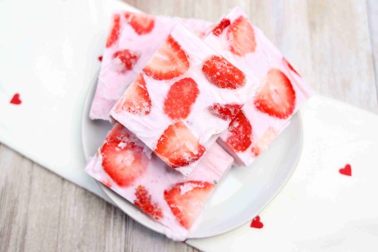 Four squares of yogurt bark with visible slices of fresh strawberries on top are stacked on a white plate. The plate sits on a white napkin decorated with small red hearts.