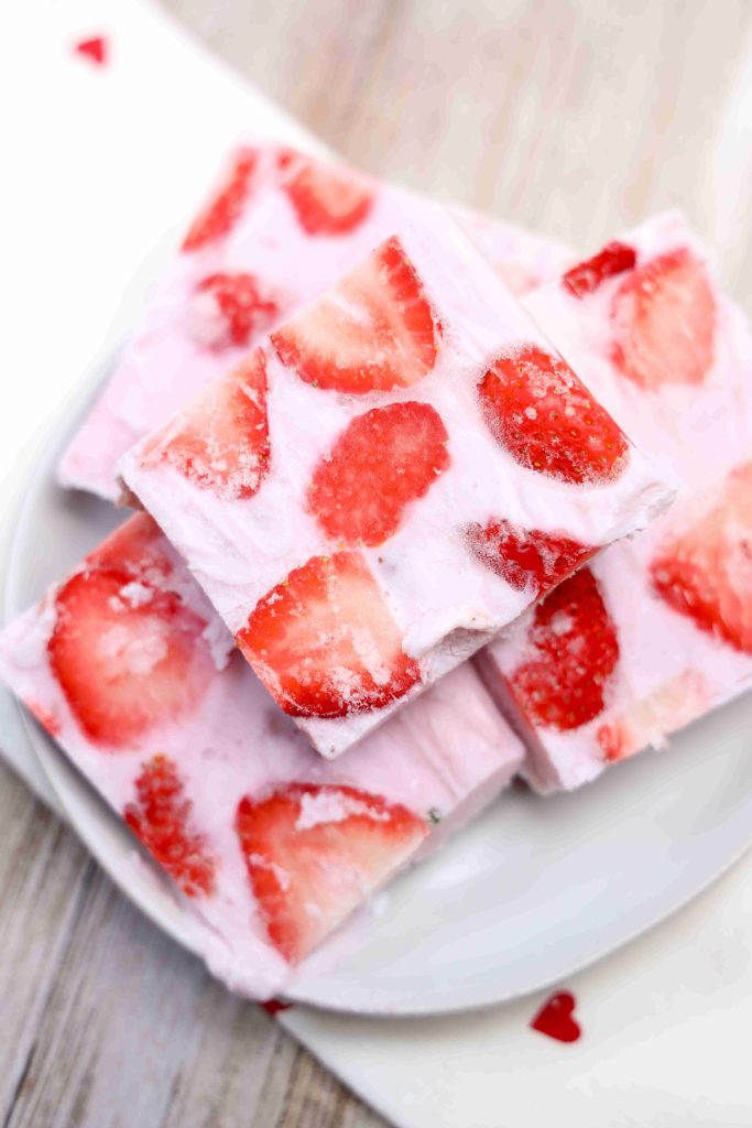 Three square pieces of yogurt bark stacked on a white plate, with slices of fresh strawberries visible in the creamy pink frozen mixture.