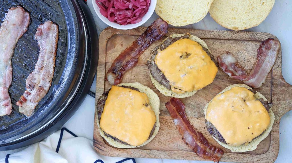 Three American Burgers with cheddar and crispy bacon strips sit on a wooden board beside a grill pan with sizzling bacon, a bowl of pickled onions, and open burger buns.