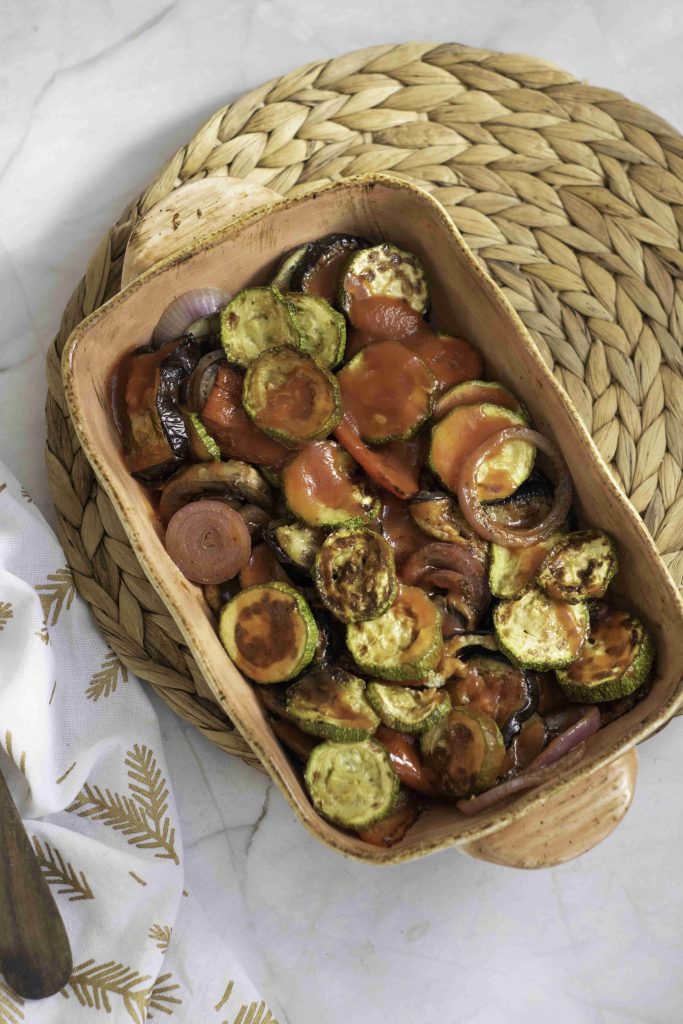 A ceramic baking dish filled with roasted vegetables, including zucchini, tomatoes, and onions, topped with tomato sauce, creates a delicious ratatouille casserole bake on a woven placemat atop a light marble surface.