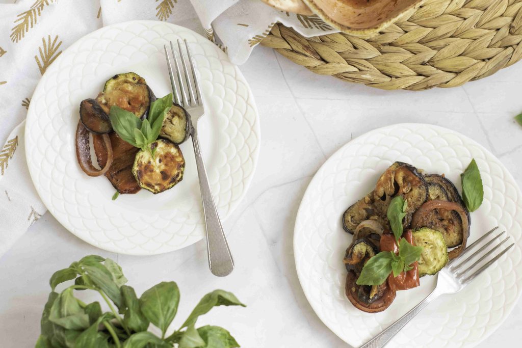 Two white plates with grilled vegetables from a ratatouille casserole bake and fresh basil, each with a fork. The plates are on a white surface next to a woven placemat and a basil plant.