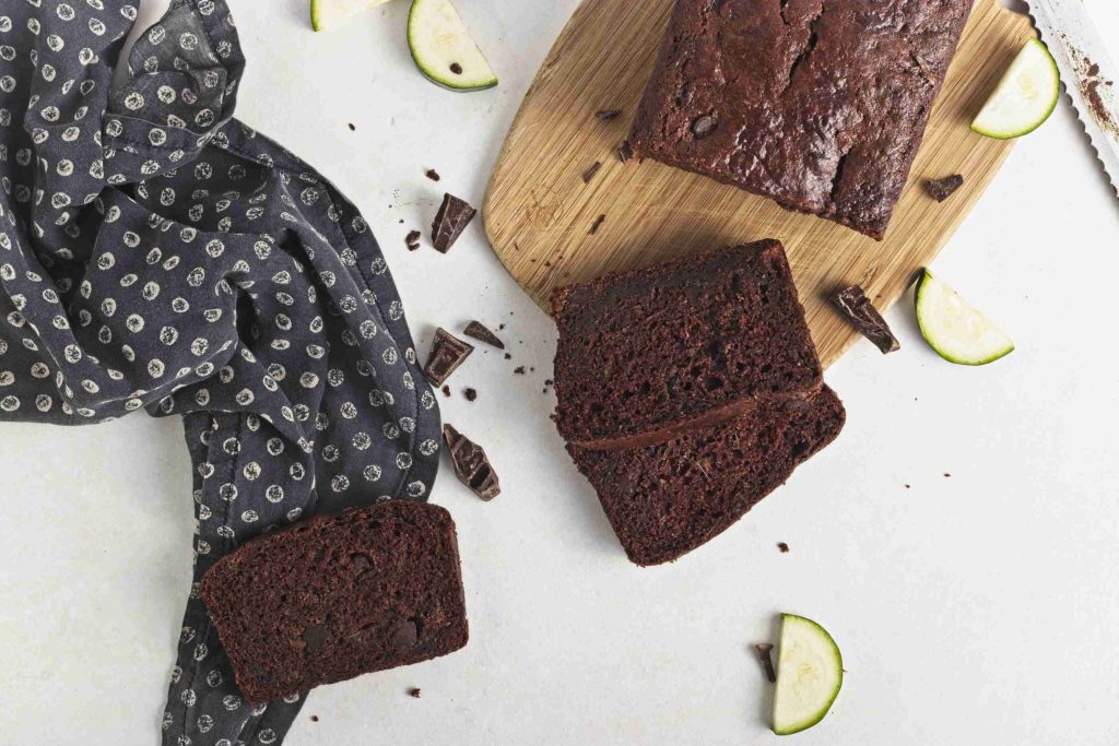 A loaf of vegan chocolate chip zucchini bread on a wooden board with two slices cut, surrounded by chocolate chunks, zucchini pieces, and a dark patterned cloth on a white surface.