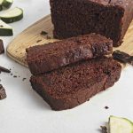 Two slices of rich vegan chocolate chip zucchini bread lie on a light surface in front of a whole loaf on a wooden board, surrounded by zucchini pieces and chunks of chocolate.