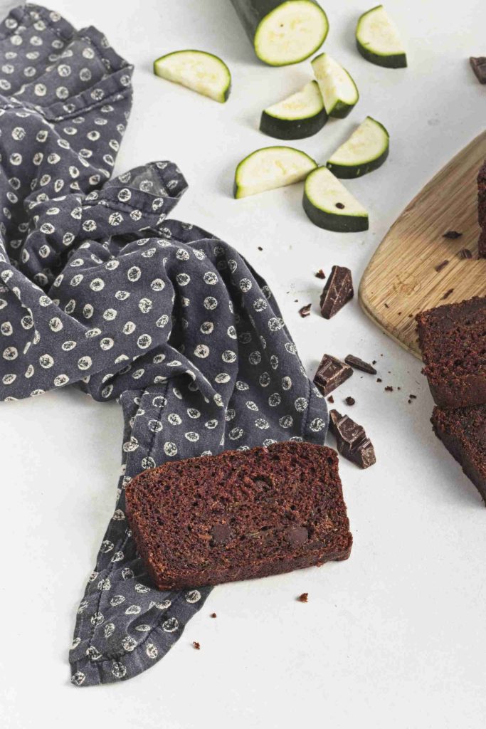A slice of vegan chocolate chip zucchini bread sits on a patterned cloth beside a wooden board with more slices, chocolate chunks, and chopped zucchini pieces on a light surface.