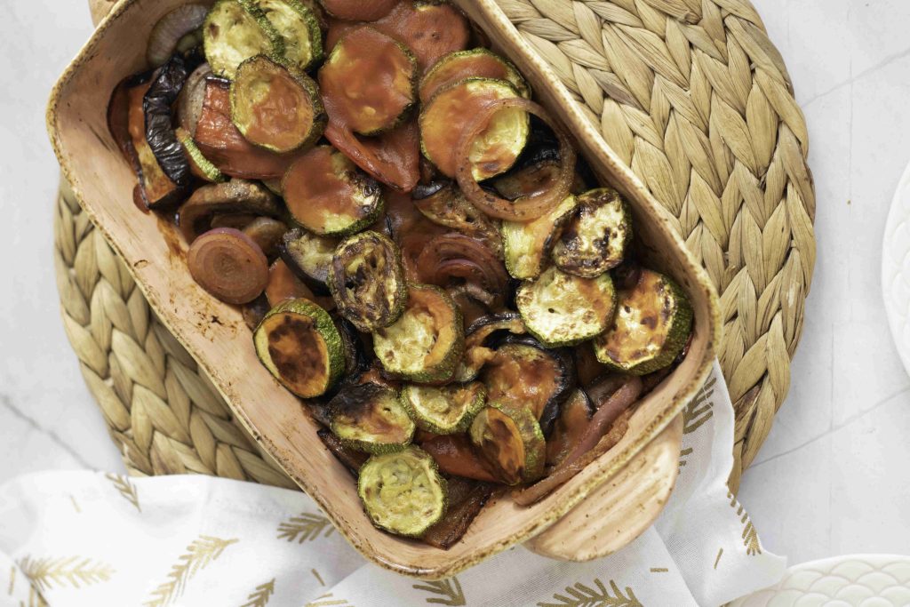 A ratatouille casserole bake filled with roasted zucchini, tomatoes, and eggplant sits on a woven placemat next to a white patterned towel on a light marble surface.