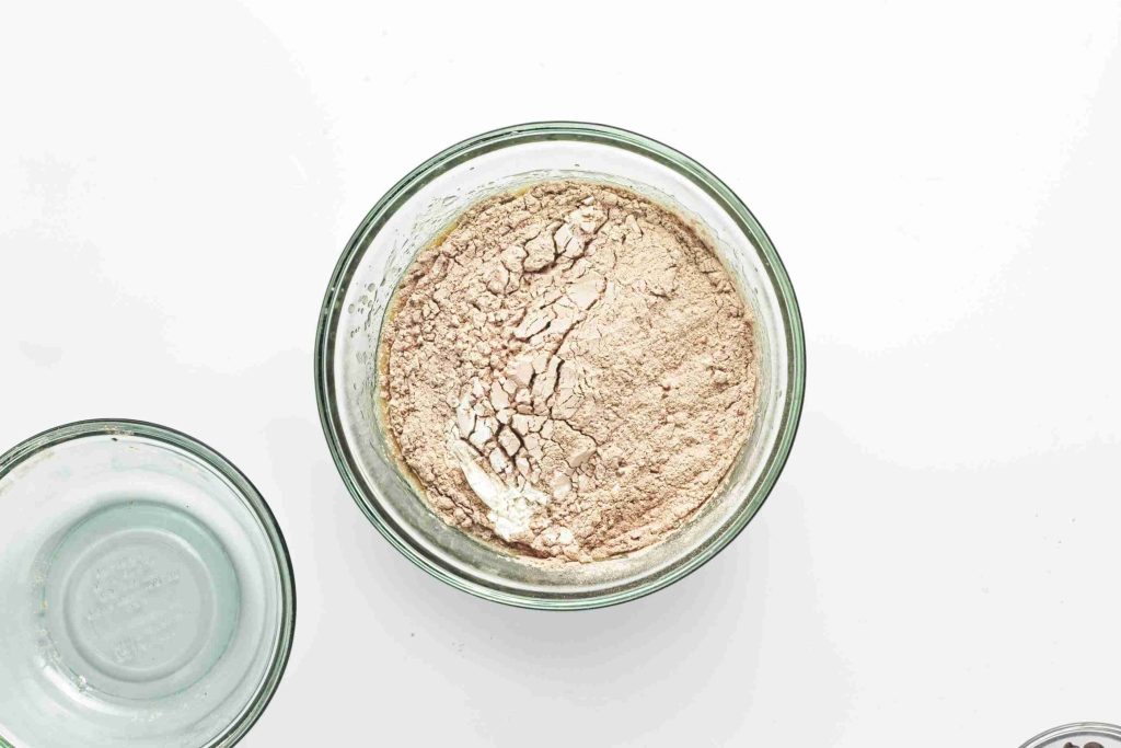 A glass bowl filled with light brown powder, possibly flour or a baking mix for vegan chocolate chip zucchini bread, sits on a white surface. Another empty glass bowl is partially visible to the left.