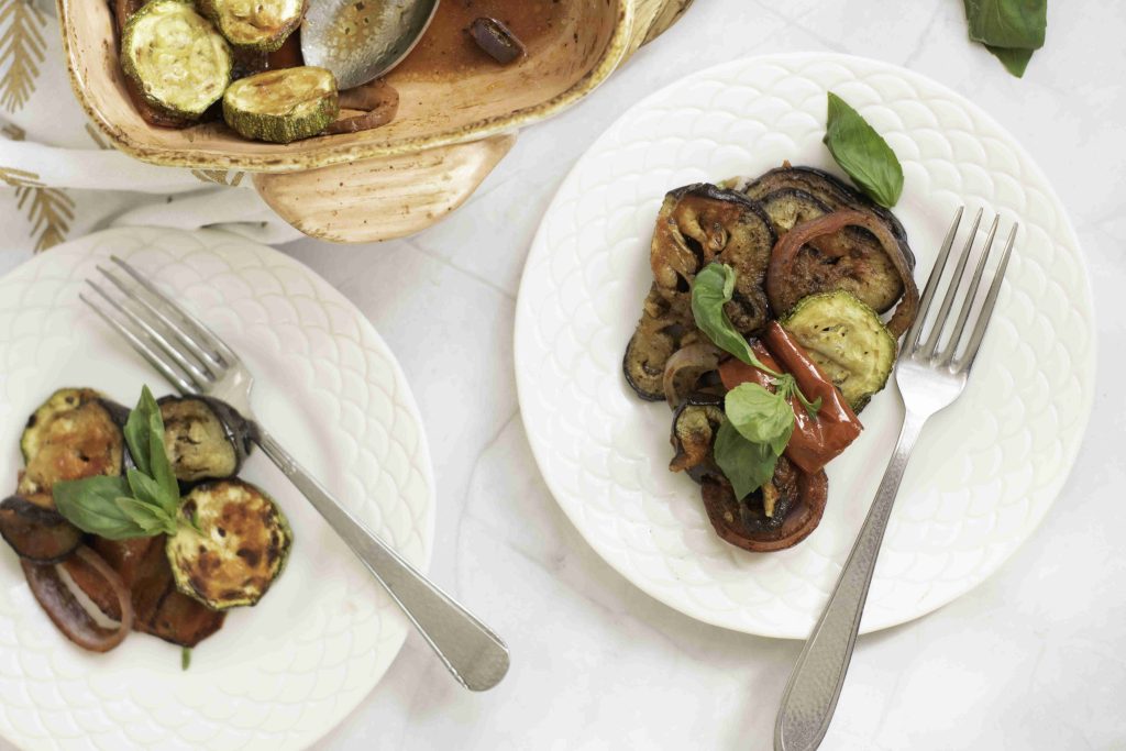 Two white plates with servings of roasted vegetables, including zucchini, tomato, and eggplant from a ratatouille casserole bake, garnished with fresh basil. Each plate has a fork beside it; a baking dish with more vegetables is partially visible above.
