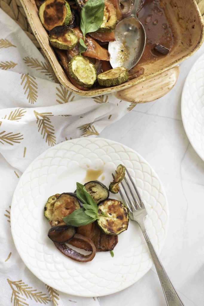 A white plate with grilled zucchini, eggplant, and tomato slices garnished with fresh basil sits next to a fork. Above, a baking dish holds a colorful ratatouille casserole bake on a patterned cloth, ready to serve.