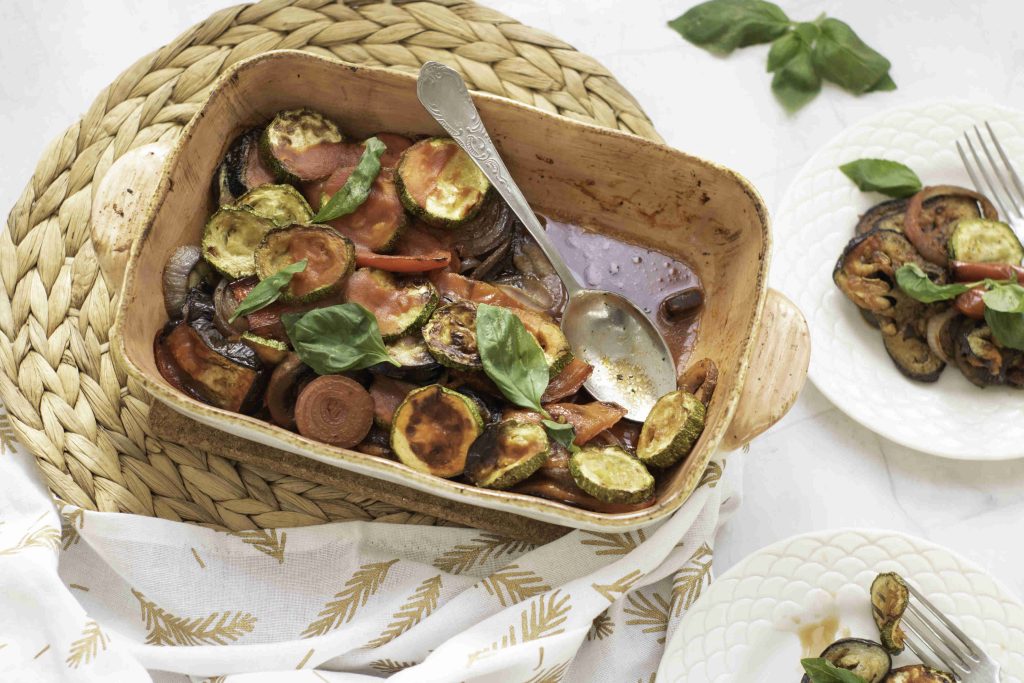 A baking dish filled with a colorful ratatouille casserole bake, featuring roasted zucchini, tomato, and eggplant, garnished with fresh basil leaves. A serving spoon rests in the dish, with two plates of the vegetables placed nearby.