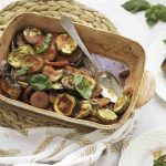 A baking dish filled with a colorful ratatouille casserole bake, featuring roasted zucchini, tomato, and eggplant, garnished with fresh basil leaves. A serving spoon rests in the dish, with two plates of the vegetables placed nearby.