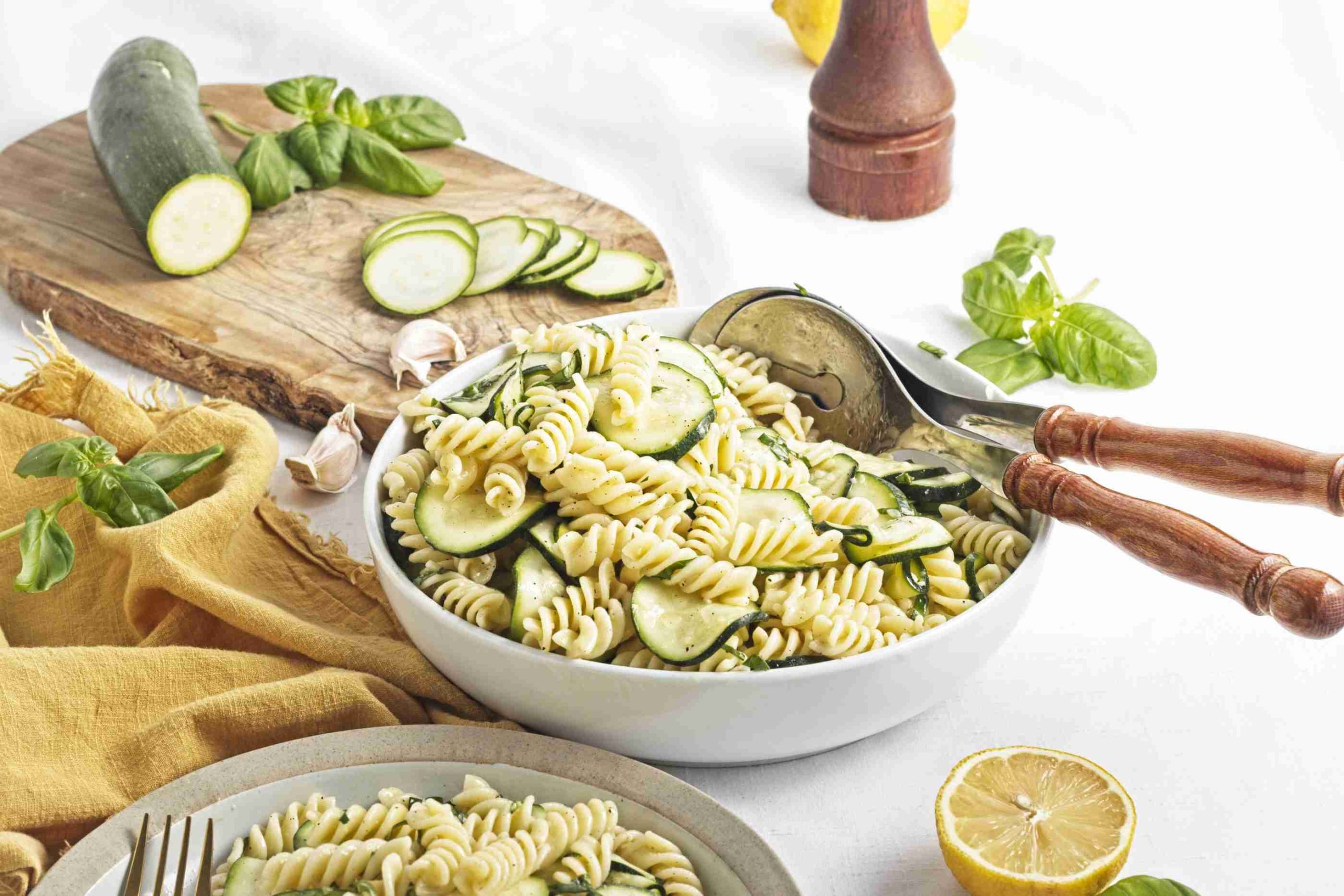 A bowl of zucchini pasta salad with rotini and fresh basil sits on a white table beside a pepper grinder, lemon halves, garlic, more sliced zucchini on a cutting board, and serving utensils resting in the bowl.