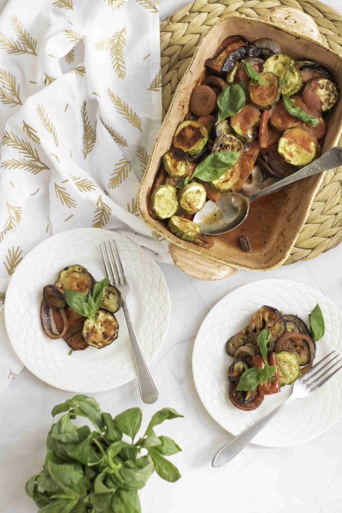 A baked ratatouille casserole with sliced zucchini, tomatoes, and eggplant is served on two white plates with forks. Fresh basil leaves garnish the plates. A patterned napkin and a potted basil plant are nearby.