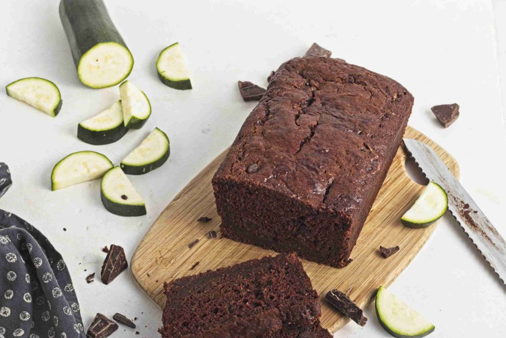A loaf of vegan chocolate chip zucchini bread sits on a wooden cutting board, with one slice cut. Sliced zucchini, chocolate chunks, and a knife are nearby on a white surface.