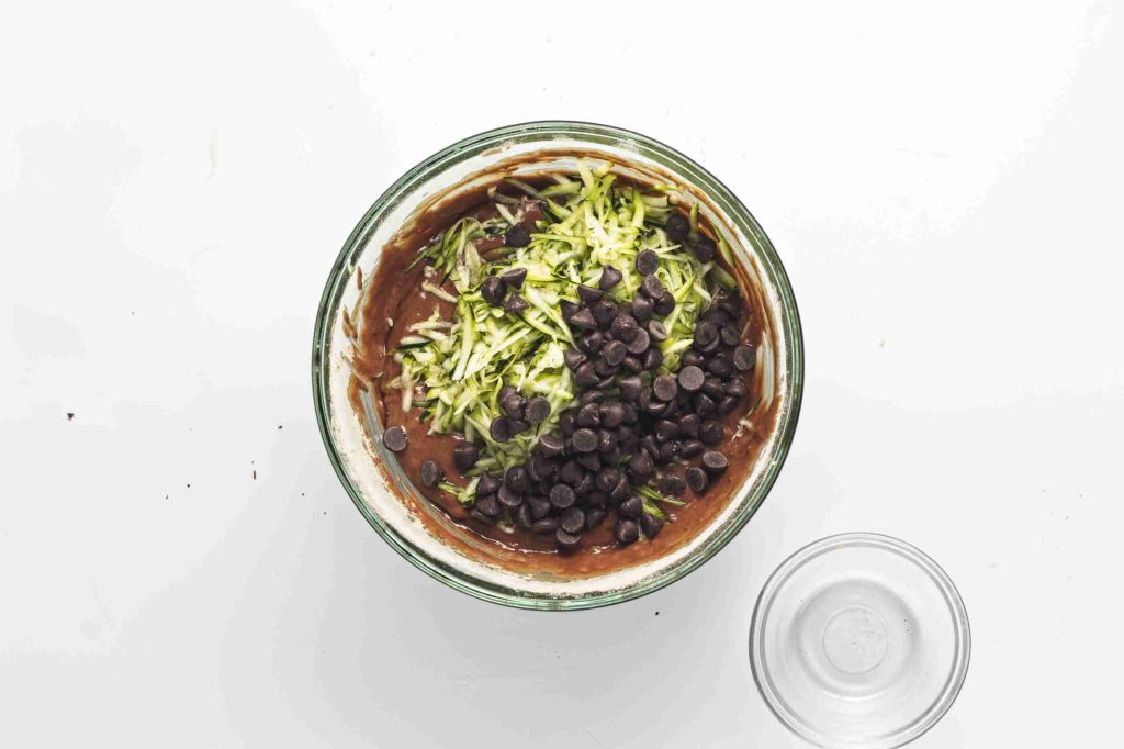 A glass bowl filled with vegan chocolate chip zucchini bread batter, shredded zucchini, and chocolate chips sits on a white surface next to an empty small glass bowl.