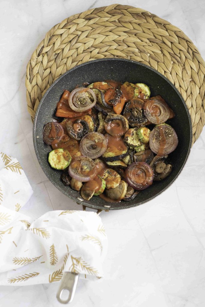 A black skillet filled with roasted vegetables, including sliced onions, zucchini, mushrooms, and carrots, sits on a woven placemat—a rustic ratatouille casserole bake. A white kitchen towel with a gold leaf pattern rests nearby on the marble surface.