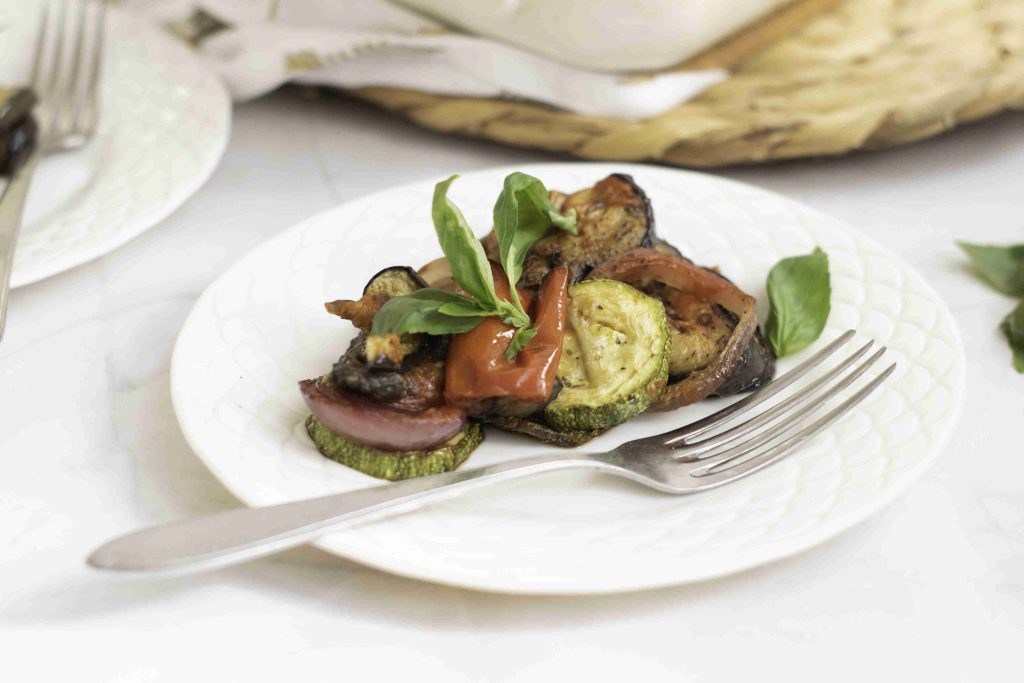 A white plate with roasted vegetables, including zucchini, red onion, and tomato, from a ratatouille casserole bake, garnished with fresh basil leaves. A silver fork rests on the plate.