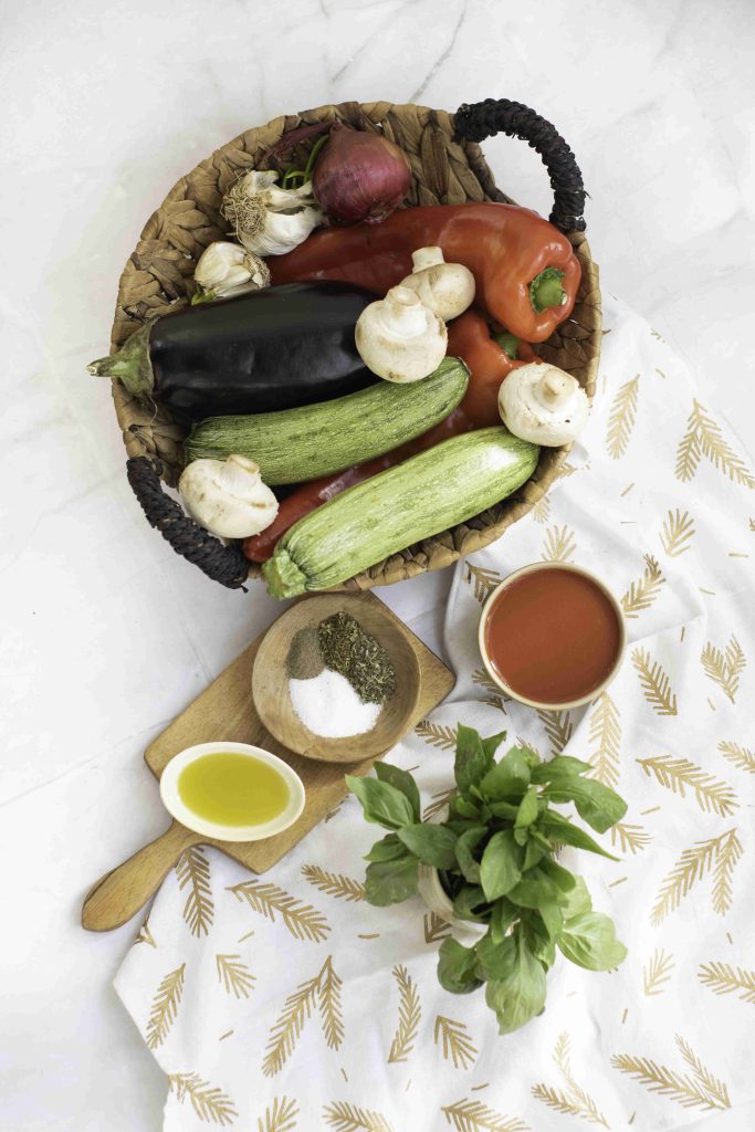 A woven basket holds vegetables perfect for a ratatouille casserole, including zucchini, eggplant, and peppers. Nearby are basil, olive oil, mixed herbs and salt, and a glass of tomato sauce to complete the flavorful bake.