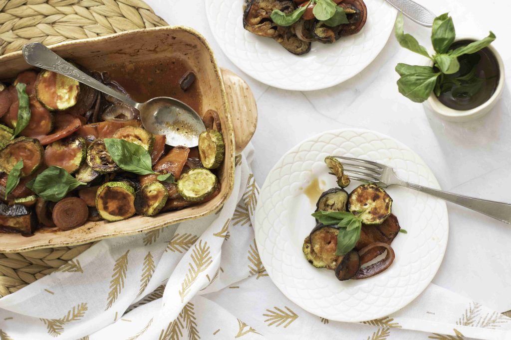 A ratatouille casserole bake with zucchini, tomatoes, and onions topped with fresh basil sits next to two white plates serving portions of the dish. A cup of basil and a patterned napkin are also on the table.