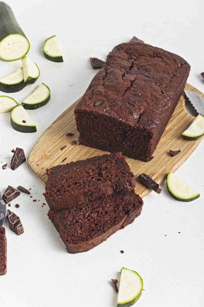 A vegan chocolate chip zucchini bread loaf rests on a wooden cutting board, with two slices cut in front. Pieces of chocolate and sliced zucchini are scattered around on a white surface.
