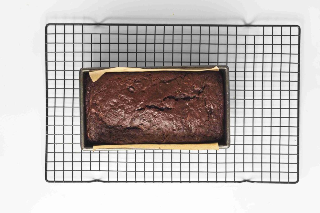 A loaf of vegan chocolate chip zucchini bread sits in a rectangular baking pan lined with parchment paper, placed on a black wire cooling rack against a white background.
