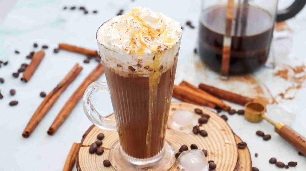 A glass mug of iced cinnamon almond milk latte topped with whipped cream and sprinkled cinnamon, surrounded by coffee beans, cinnamon sticks, ice cubes, and a French press in the background.